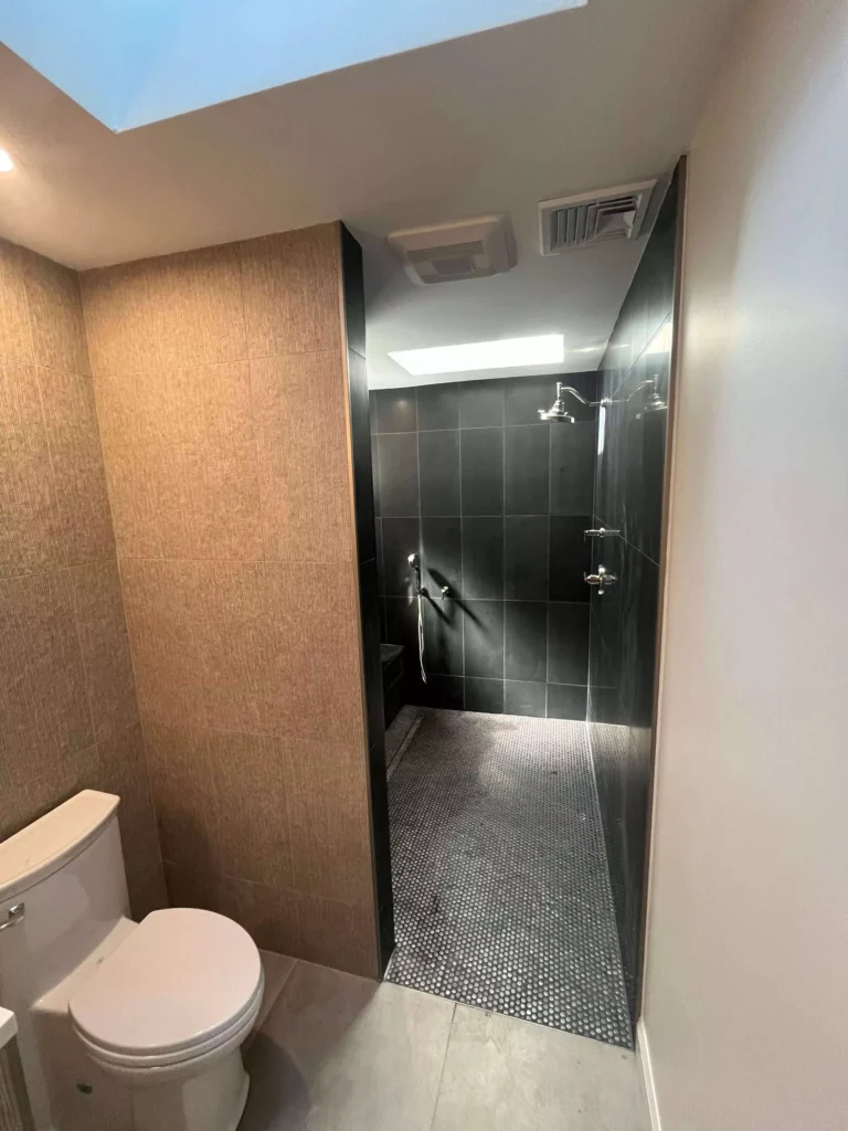 Modern bathroom remodel featuring a white toilet, light brown textured wall, and a dark tiled shower with black penny floor.