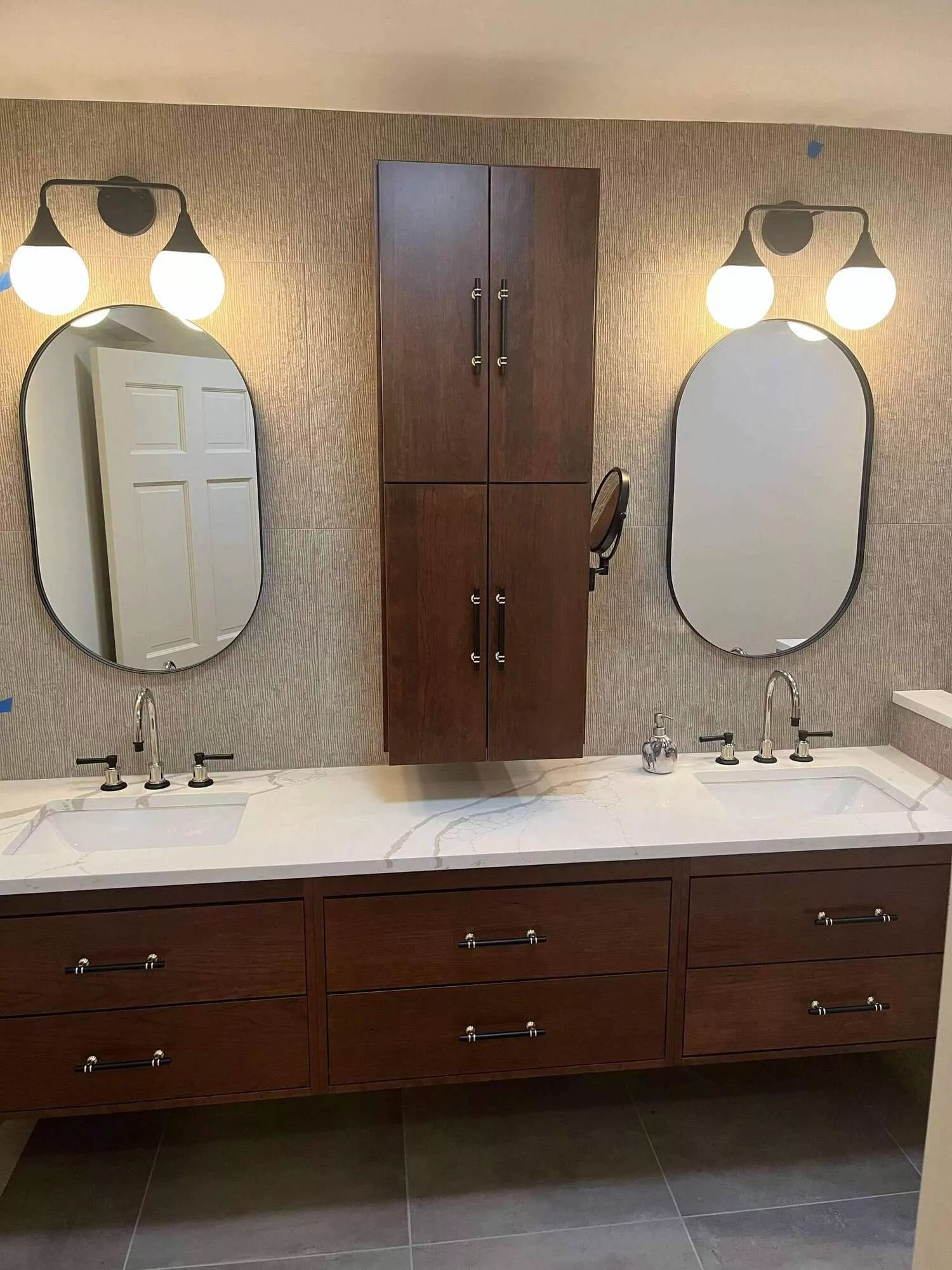 Modern bathroom double vanity with dark wood floating cabinets, white marble counter, two oval mirrors, and wall lights.