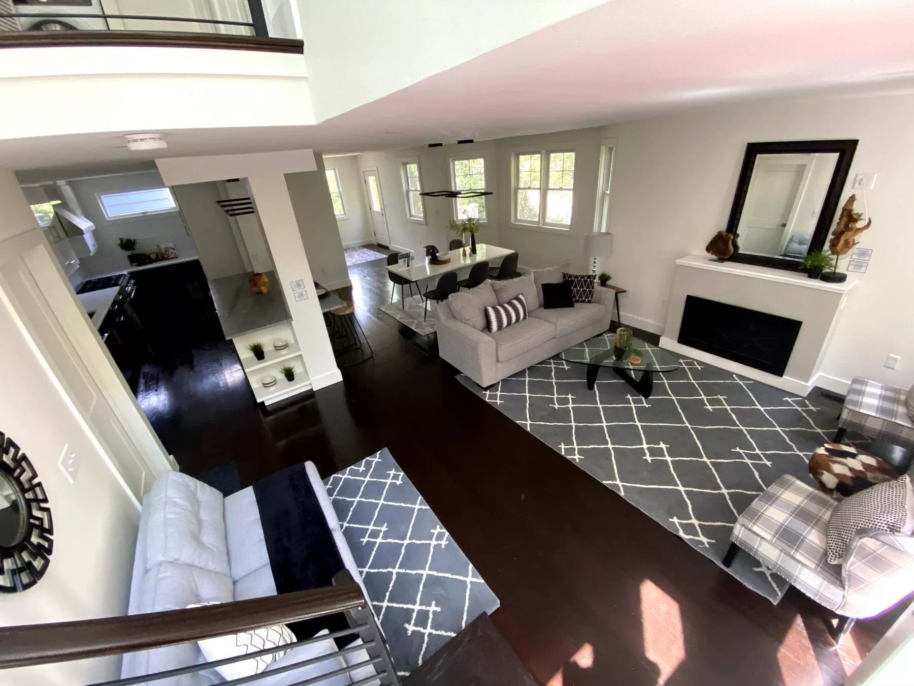 Bright, open-concept remodeled living space with grey sofa, patterned rug, fireplace, dining area, and dark kitchen.