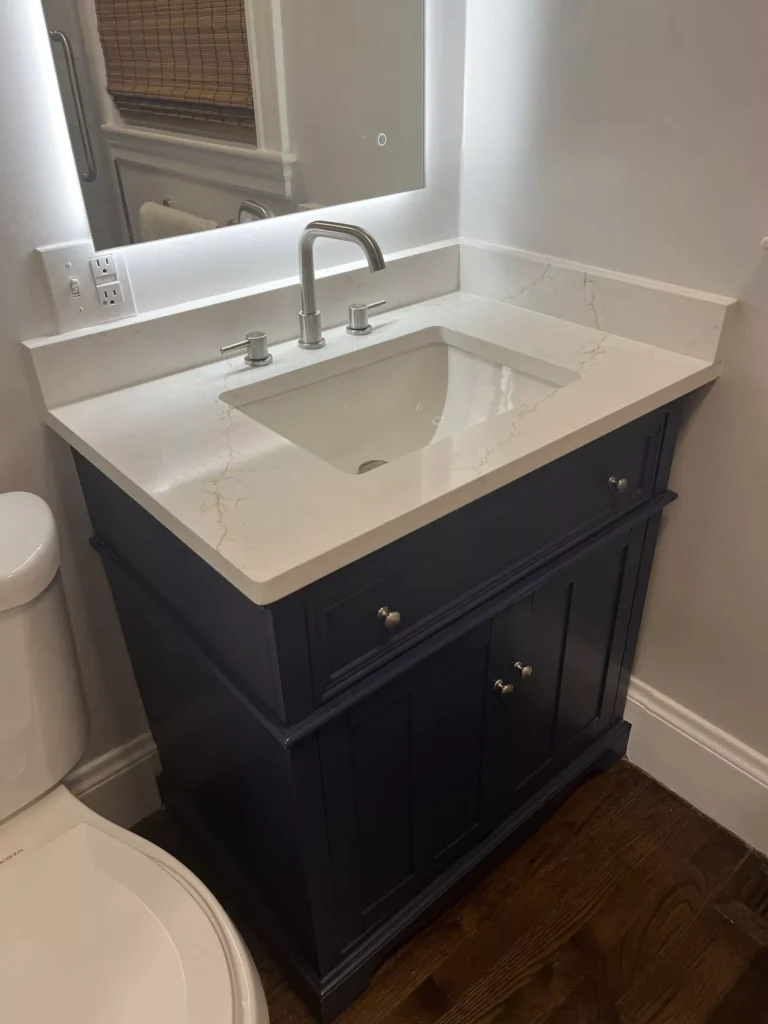 Dark blue bathroom vanity with white quartz countertop, undermount sink, brushed nickel faucet, and backlit mirror. Modern...