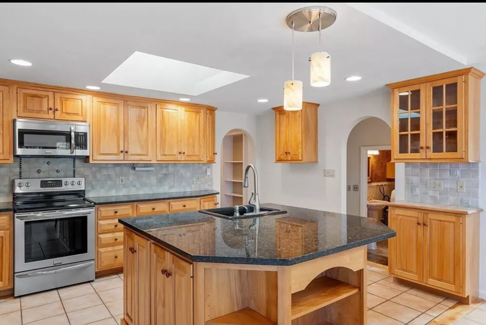 Bright kitchen with light wood cabinets, stainless steel appliances, black granite countertops, island with sink, pendant ...