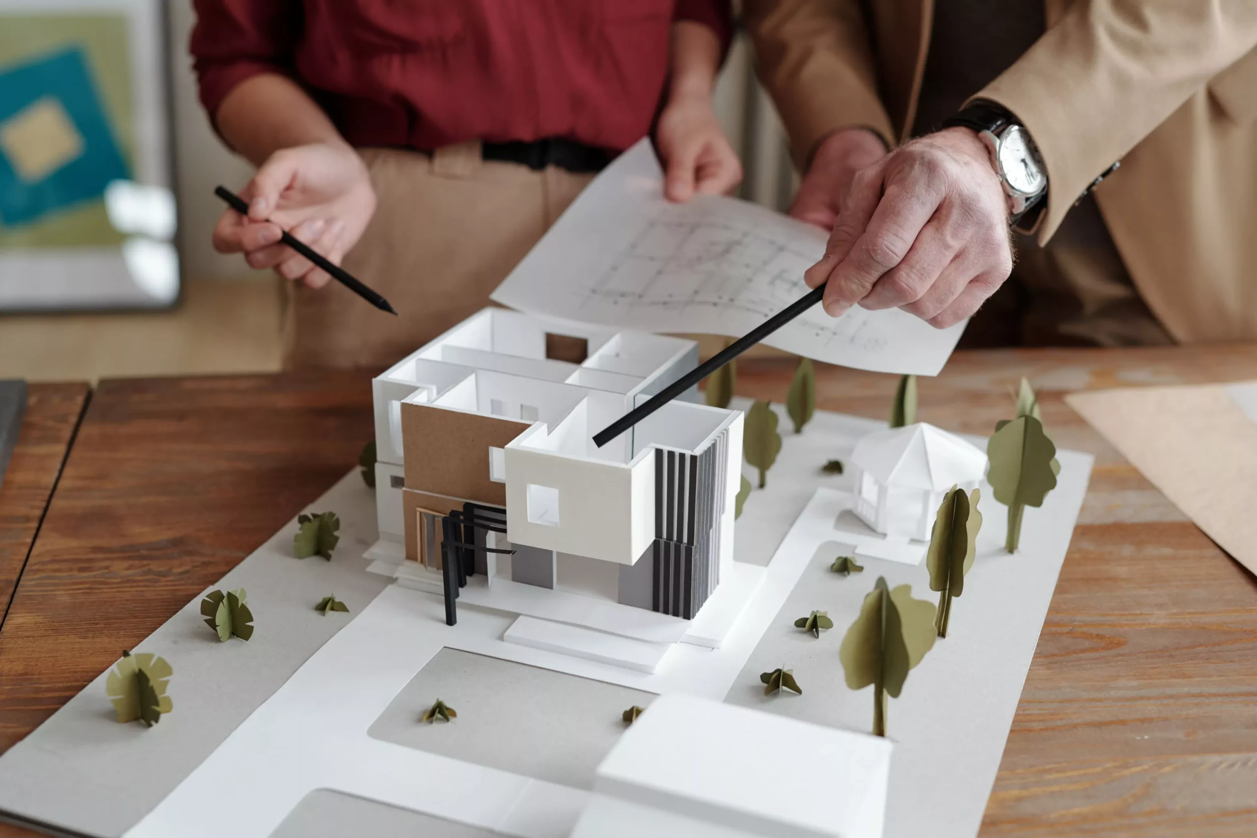 Two pairs of hands, one holding blueprints, point with pencils at a modern white and brown house model with paper trees on...