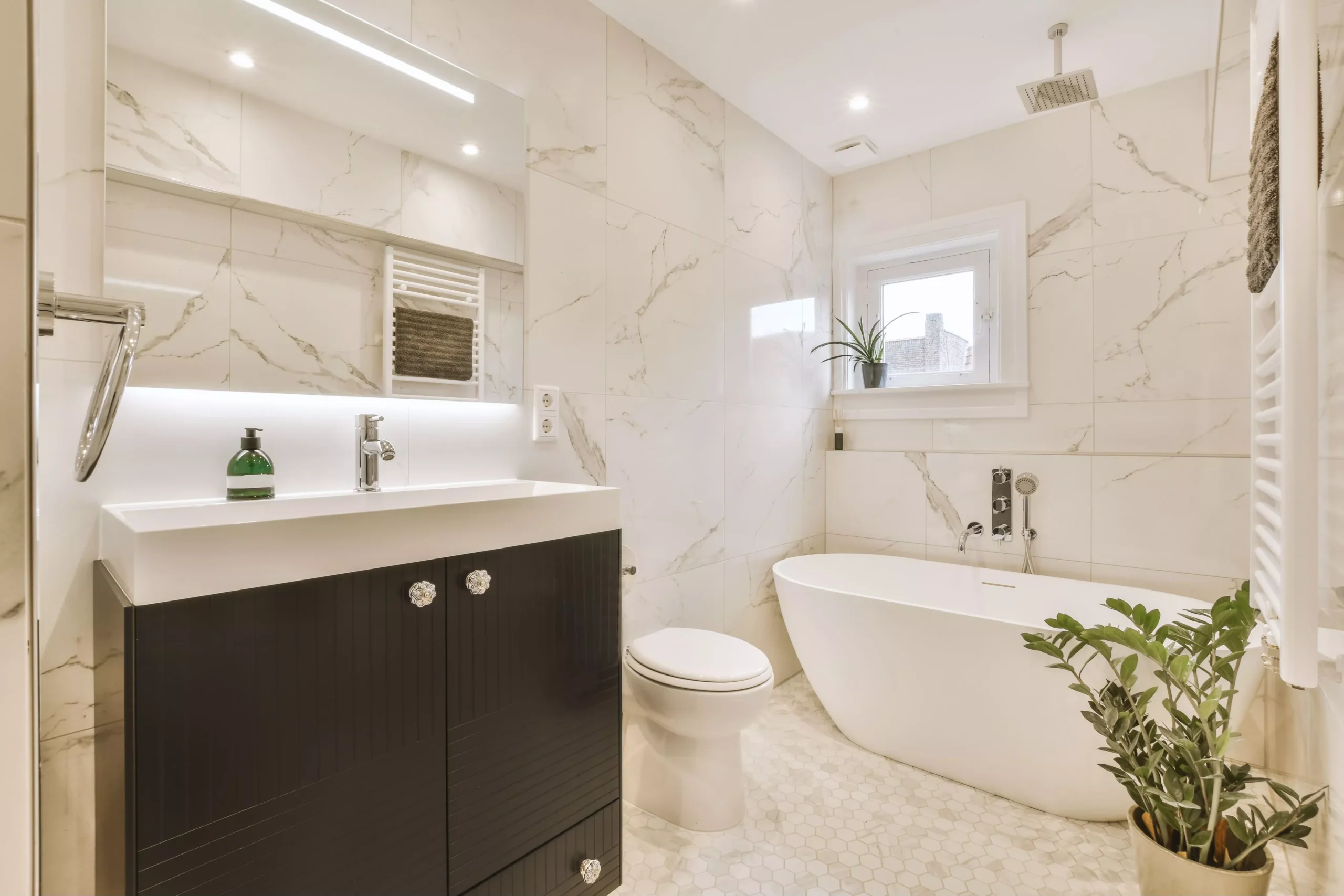 Elegant modern bathroom with white marble-tiled walls, dark brown vanity, sink, mirror, freestanding tub, and green plants.