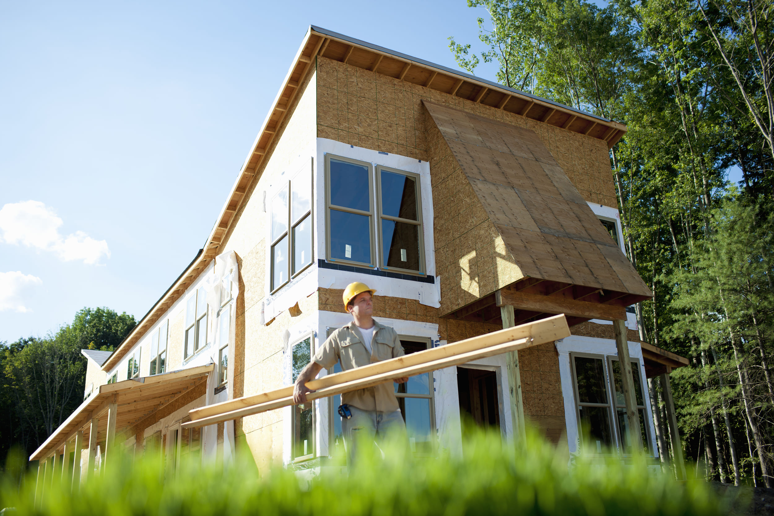Construction worker in yellow hard hat carrying wood planks in front of a house under construction with green trees and bl...