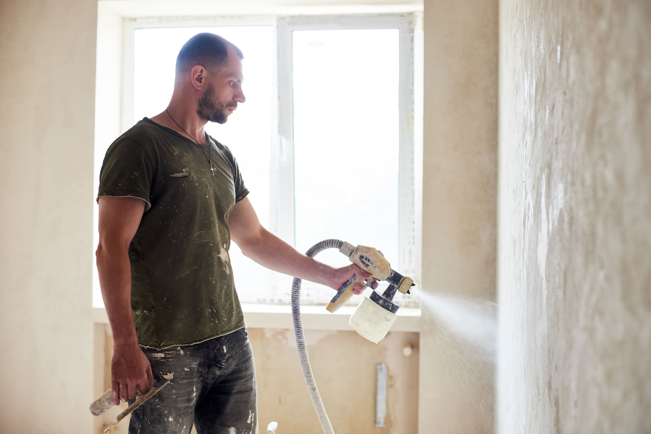 Bearded man in dirty green shirt painting a textured beige wall with a paint sprayer during home renovation.